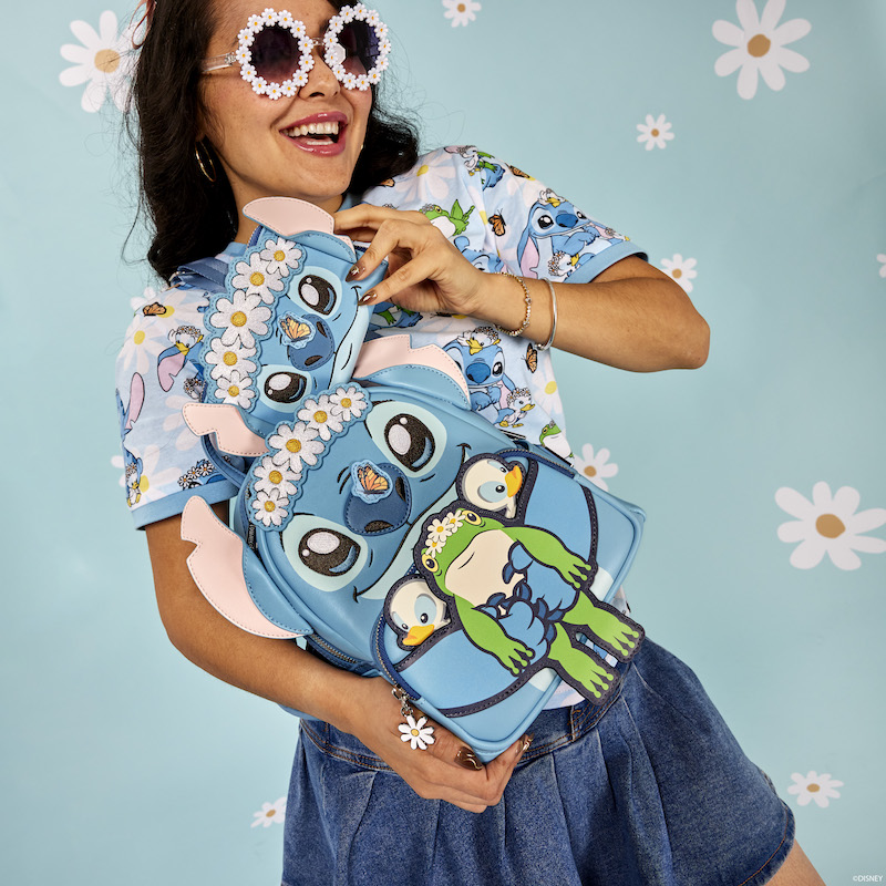 Woman wearing daisy sunglasses, wearing the Stitch Springtime Daisy unisex tee and a darker blue skirt, holding the Loungefly Stitch Springtime Daisy Cosplay Mini Backpack and standing against a light blue background with daisies on it 